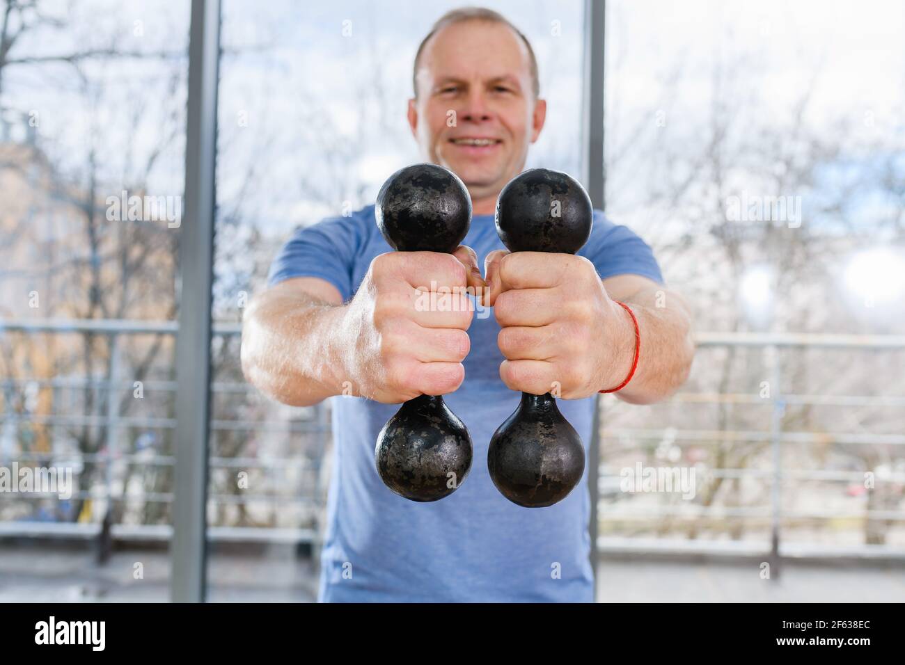 Fit aged smiling man hold two dumbbells close to camera near light gym window, healthy livestyle, workout, muscles training Stock Photo