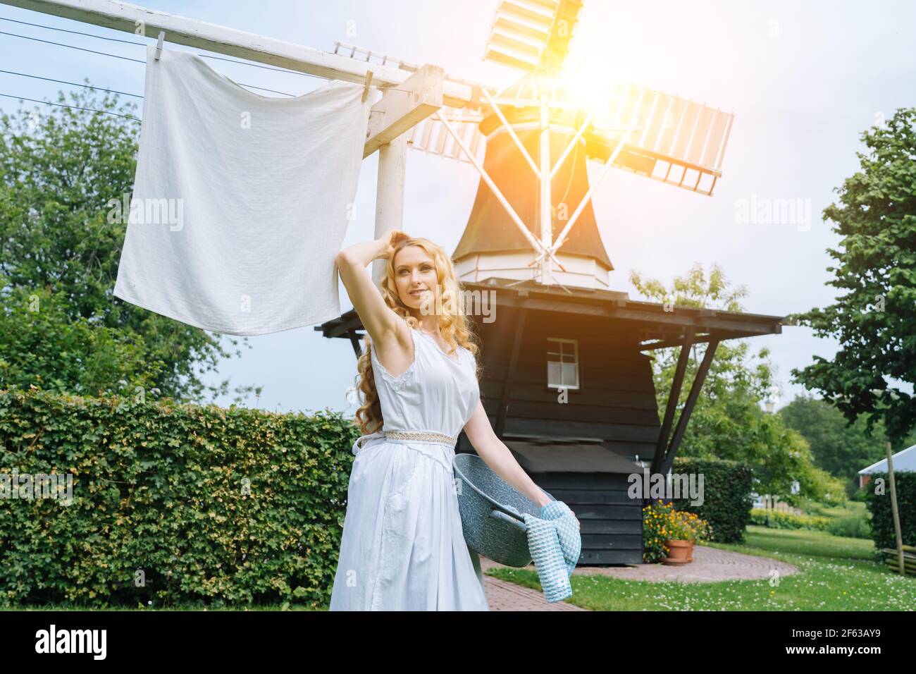 Woman dressed on traditional Dutch dress wooden shoes yellow clogs hanging Clothes on clothesline outside. laundry drying outdoor Netherlands windmill background on sun windy day. retro vintage style  Stock Photo