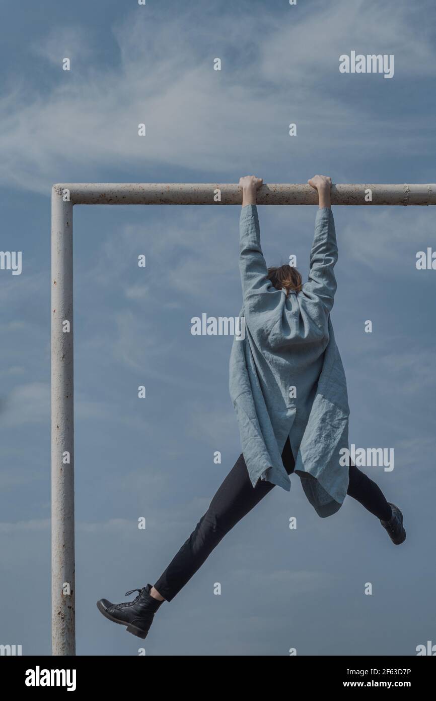 Isolated woman in black pants, aqua cardigan hanging in the air holding a metal bar with hands, legs wide apart, with beautiful sky on the background Stock Photo