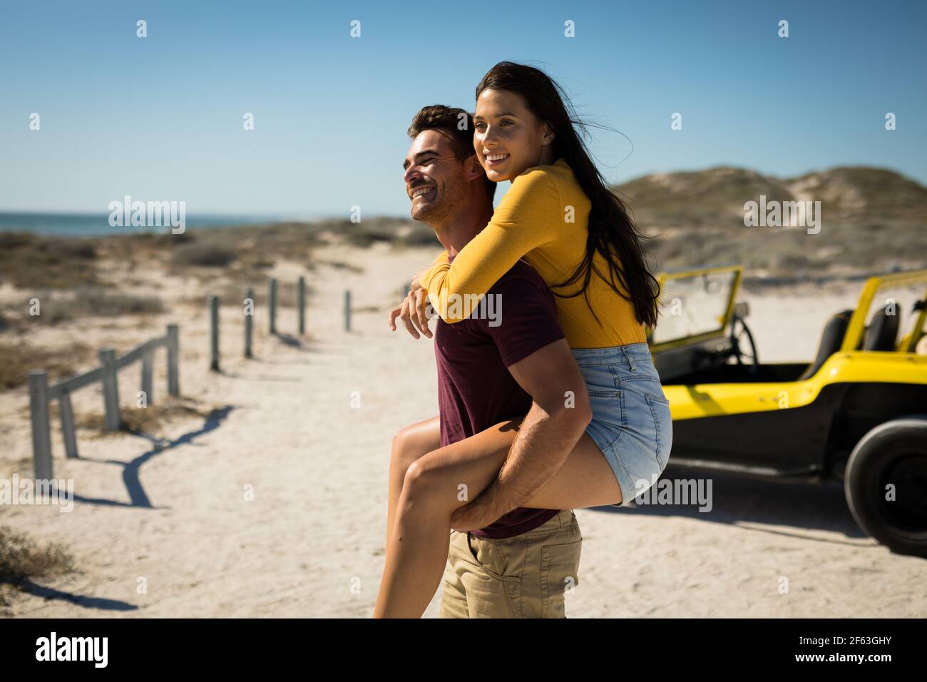Happy caucasian couple next to beach buggy by the sea piggybacking Stock Photo