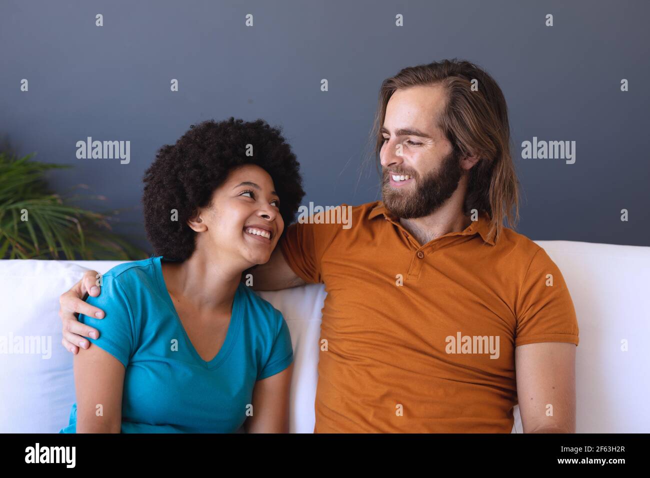 Happy diverse couple sitting on sofa embracing and smiling Stock Photo