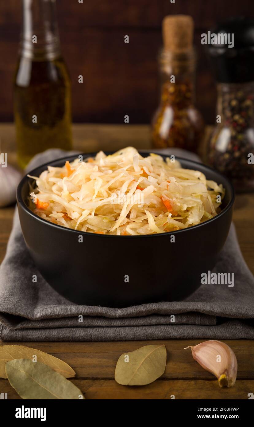 Black bowl with sour sauerkraut on gray napkin on wooden brown background close up. Next to is garlic, bay leaf, spicy, olive oil Stock Photo