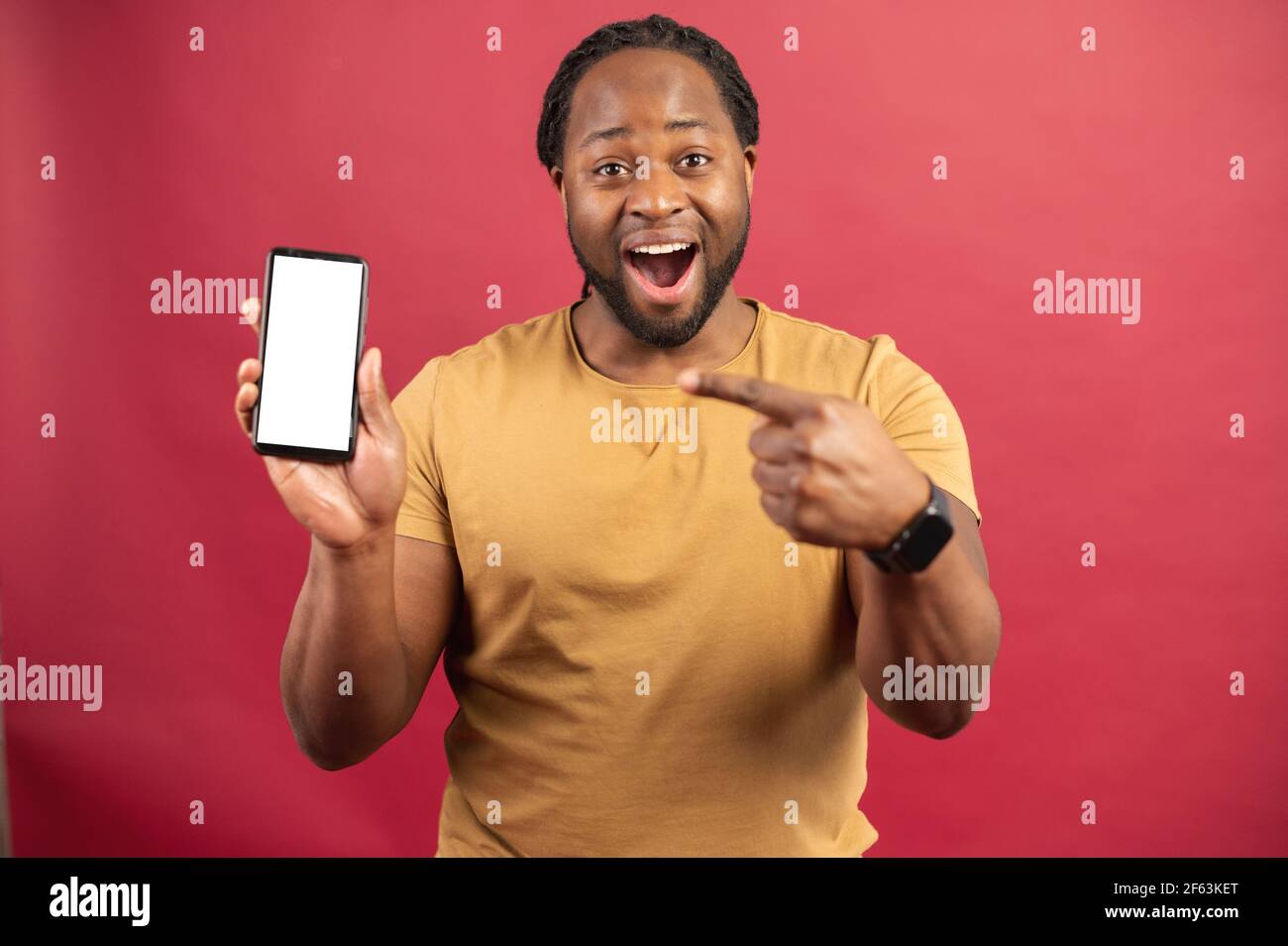 Surprised African-American man holds smartphone in a hand and points finger at empty phone screen, astonishment black guy presenting best deal, advertising new mobile app, isolated on red Stock Photo