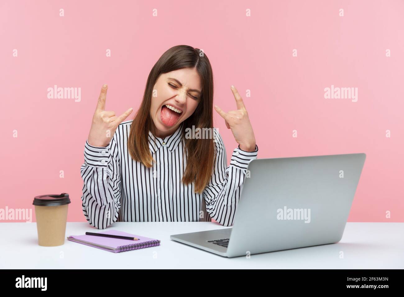 Positive naughty woman blogger showing rock and roll finger gesture looking at laptop display, chatting web camera sitting at workplace. Indoor studio Stock Photo