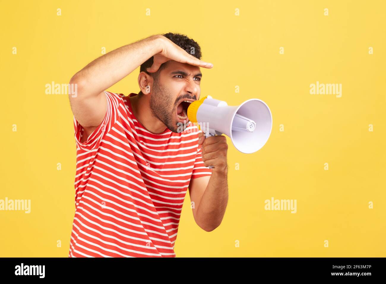 Displeased nervous man with beard in striped t-shirt loudly screaming at bullhorn megaphone holding hand on forehead and looking at distance, swearing Stock Photo