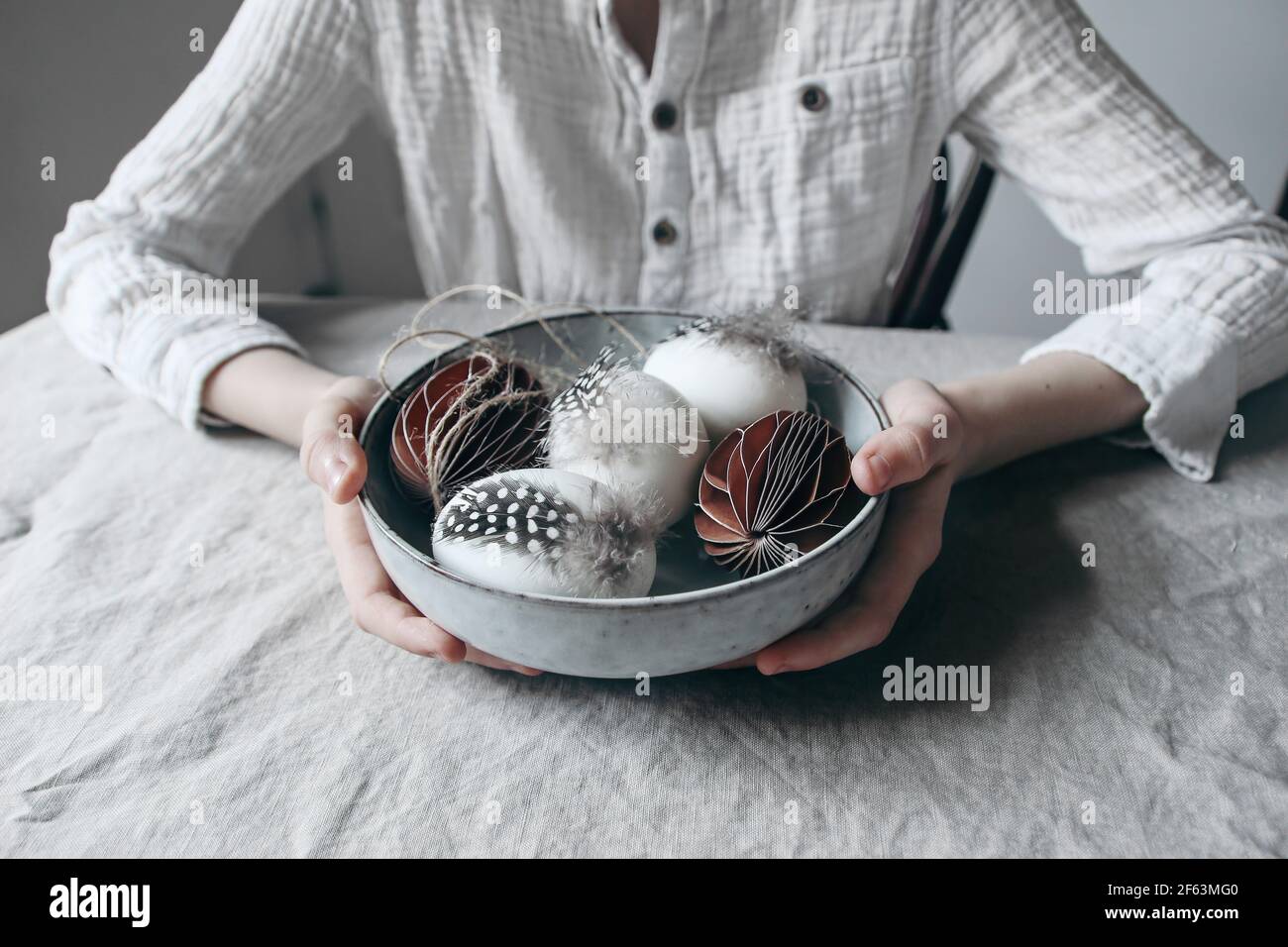 Close-up child hands in white cotton shirt holding ceramic bowl with decorative Easter eggs and paper ornaments. Moody spring lifestyle composition Stock Photo