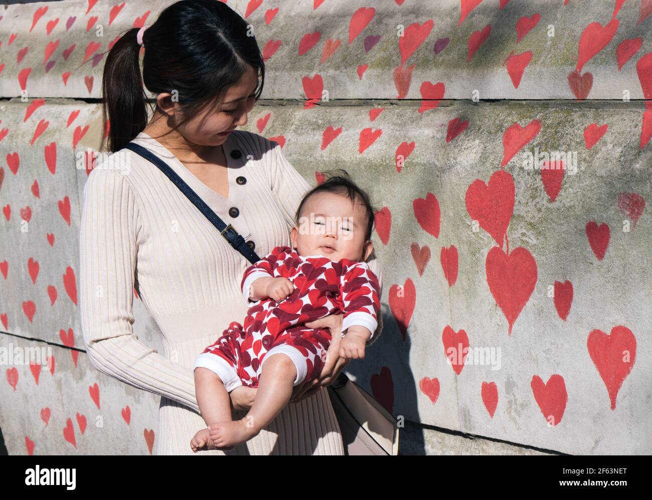 St Thomas's Hospital, London, UK. 29th March 2021. The bereaved and volunteers paint hearts for each person who has died of Covid. A woman holds a baby dressed in a heart patterned baby grow in front of the wall. Credit: Denise Laura Baker/Alamy Live News Stock Photo