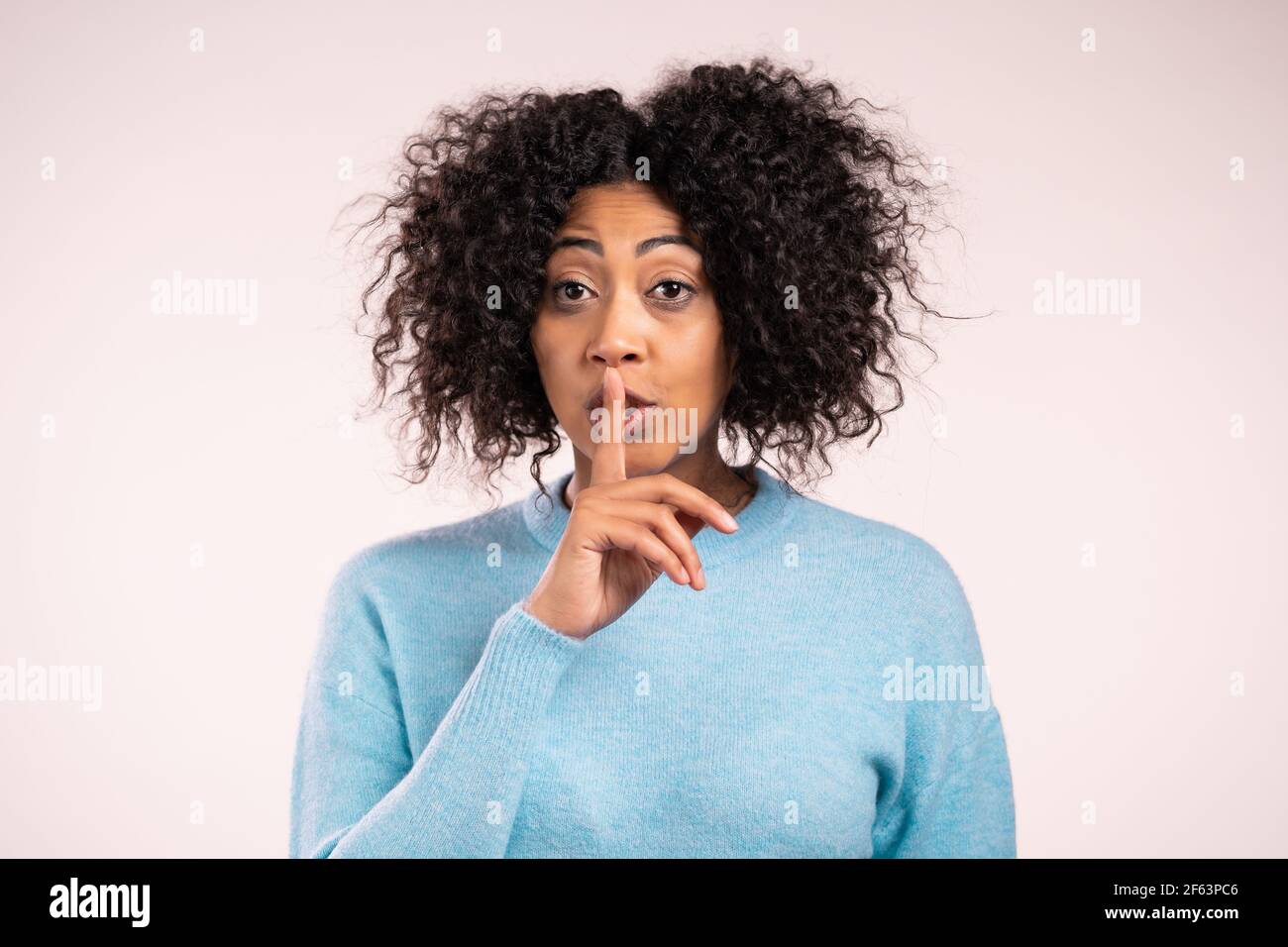 Smiling african woman with curly hair holding finger on her lips over white background. Gesture of shhh, secret, silence. Close up. Stock Photo