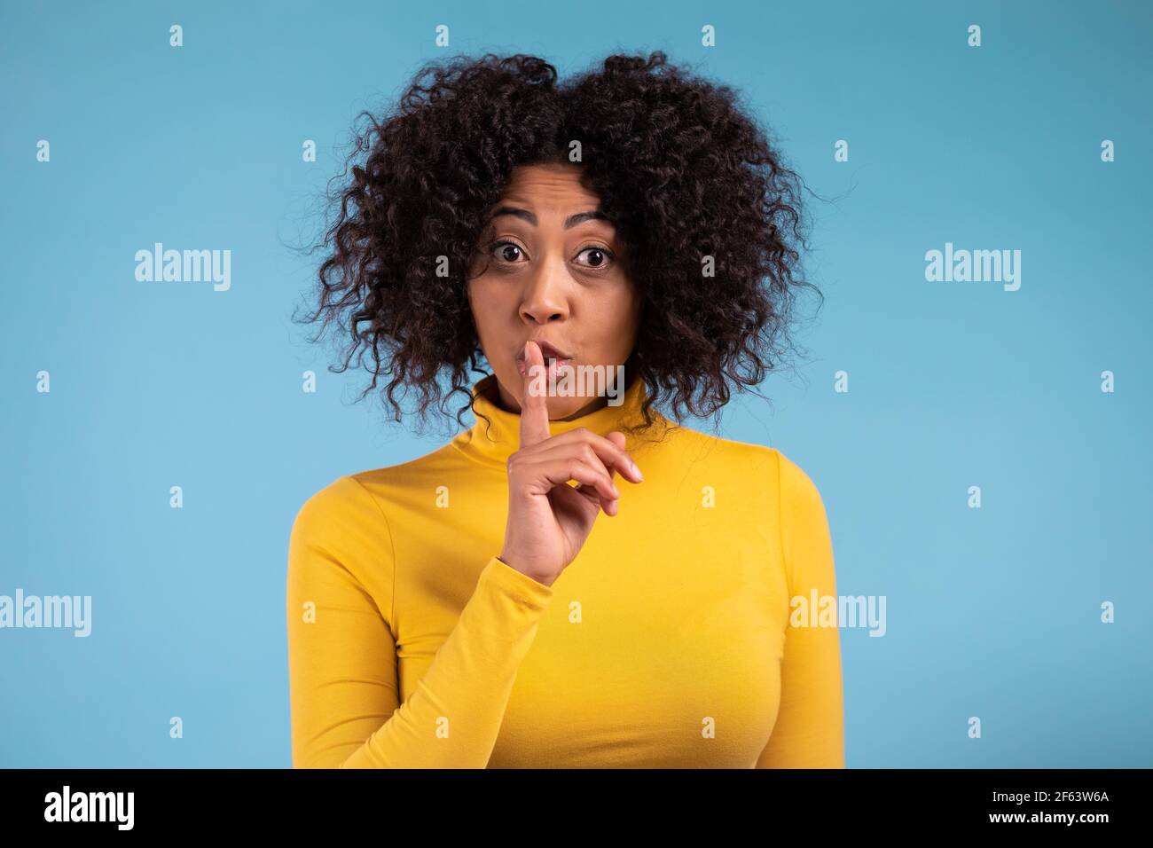 Smiling african woman with curly hair holding finger on her lips over blue background. Gesture of shhh, secret, silence. Close up Stock Photo