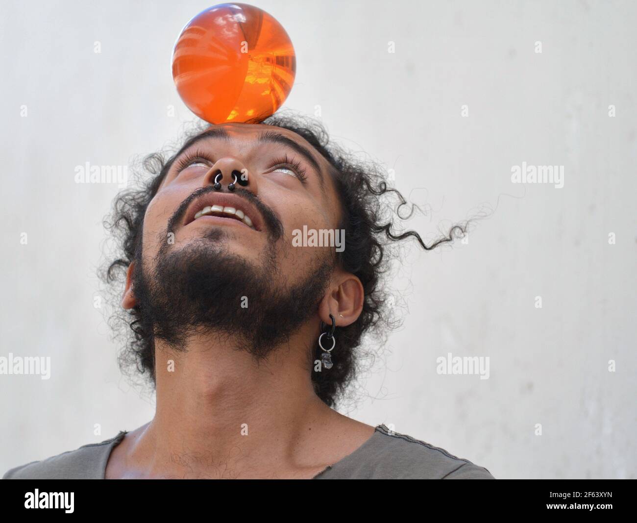 Handsome young Mexican man with septum nose jewellery balances an orange plastic ball on his forehead. Stock Photo
