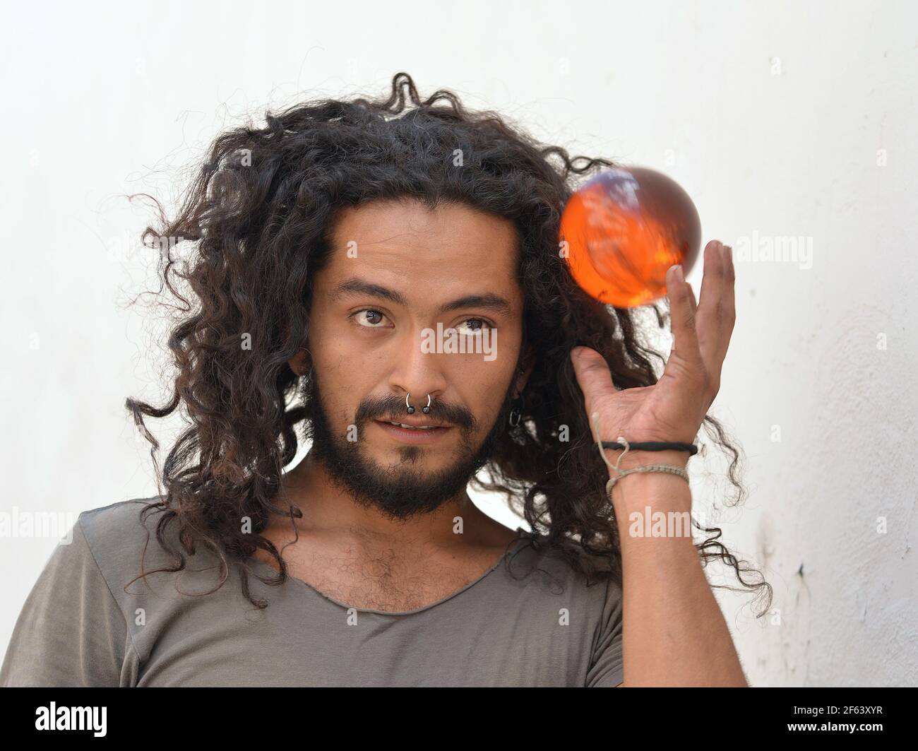 Handsome bearded young Mexican man with long natural curls and septum nose jewelry catches an orange plastic ball with his left hand. Stock Photo