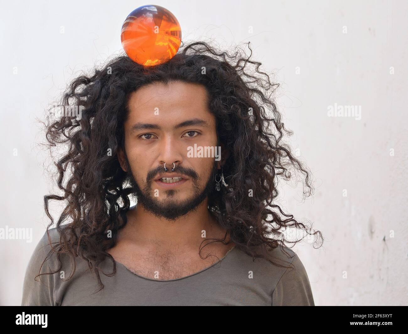 Handsome bearded young Mexican man with long natural curls and septum nose ring balances an orange plastic ball on the top of his head. Stock Photo