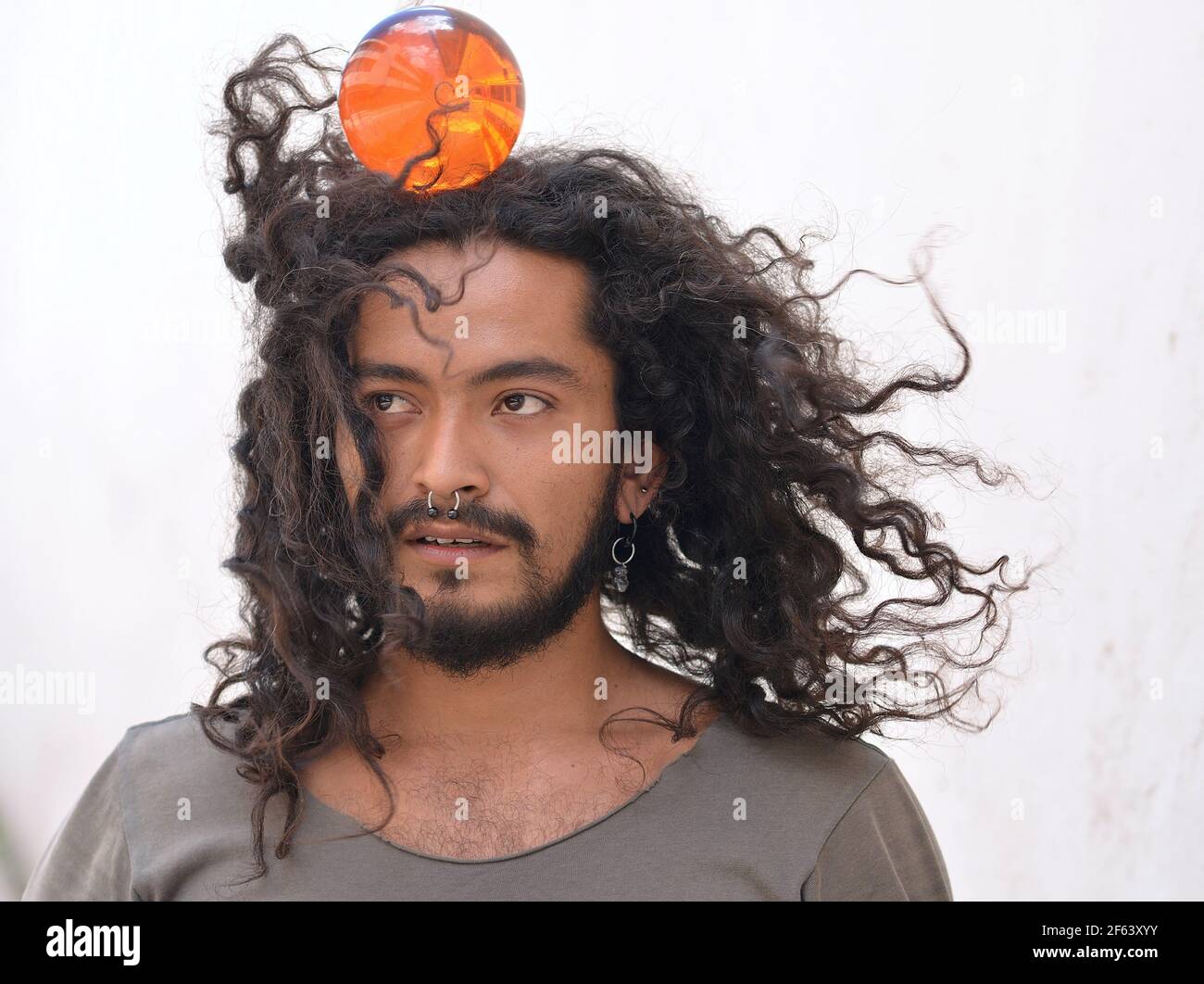 Bearded tense young Mexican man with long natural curls and septum nose ring balances an orange plastic ball on the top of his head. Stock Photo