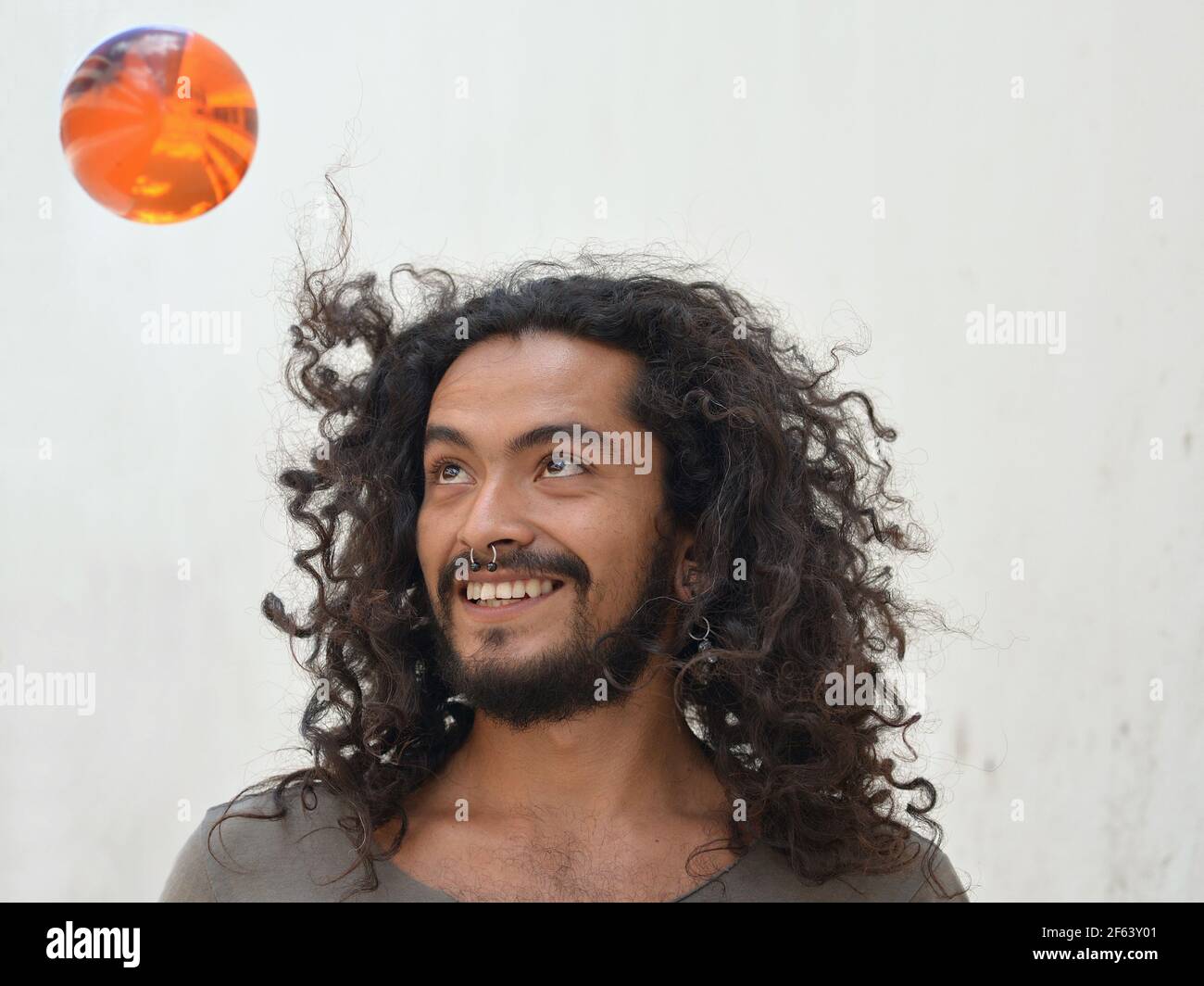 Joyful positive young Mexican man with long natural curls and septum nose jewelry plays cheerfully with an orange plastic ball. Stock Photo