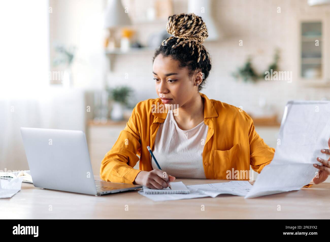 Work from home. Concentrated clever african american woman, student or designer, wearing stylish casual clothes, studying or working remotely, using laptop, watching and examines blueprints Stock Photo