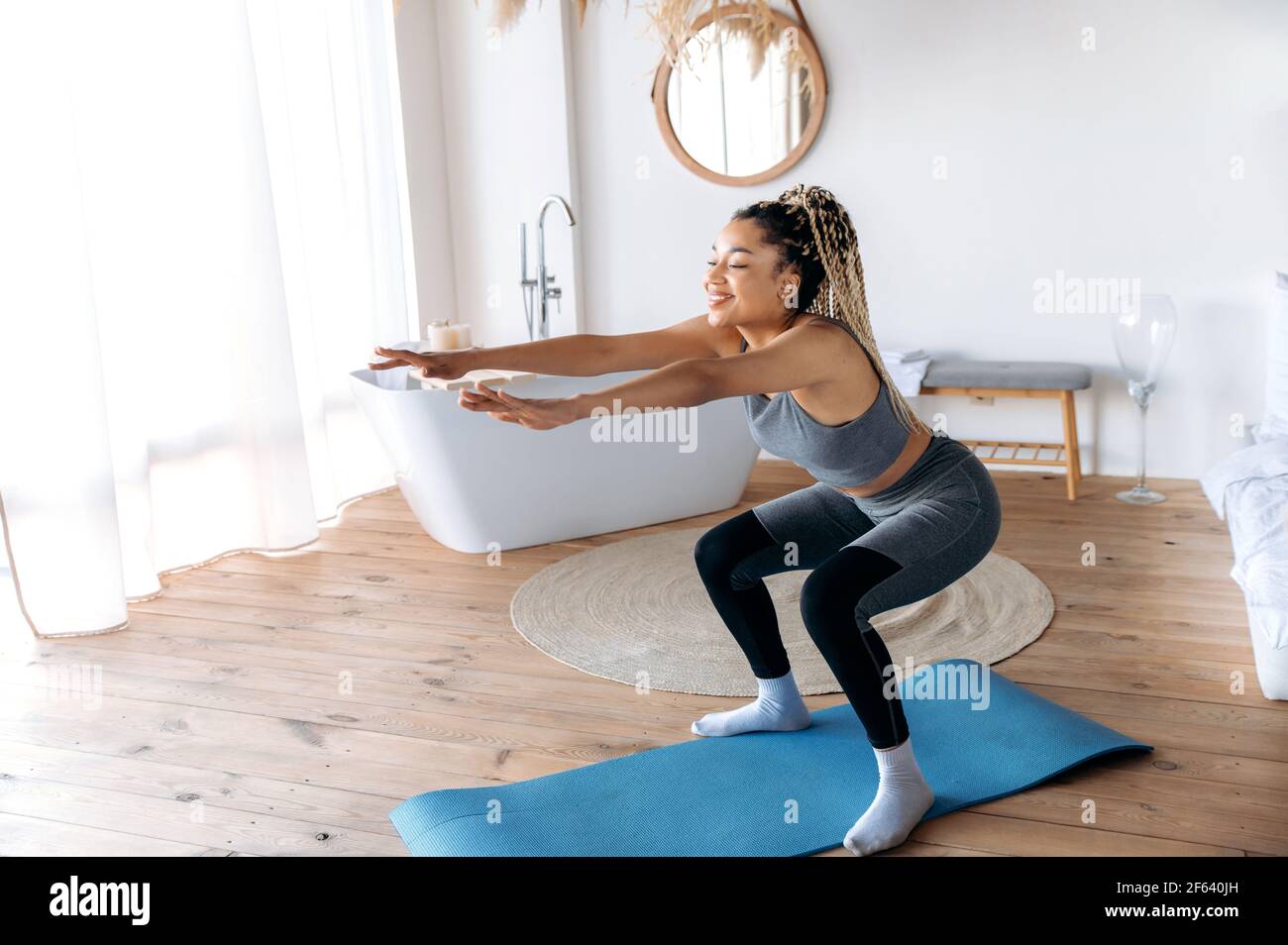 Healthy and sporty lifestyle. Attractive young African American slim sporty girl with dreadlocks care her health, does sports at home on fitness mat, does sit-ups with her arms outstretched, smiles Stock Photo