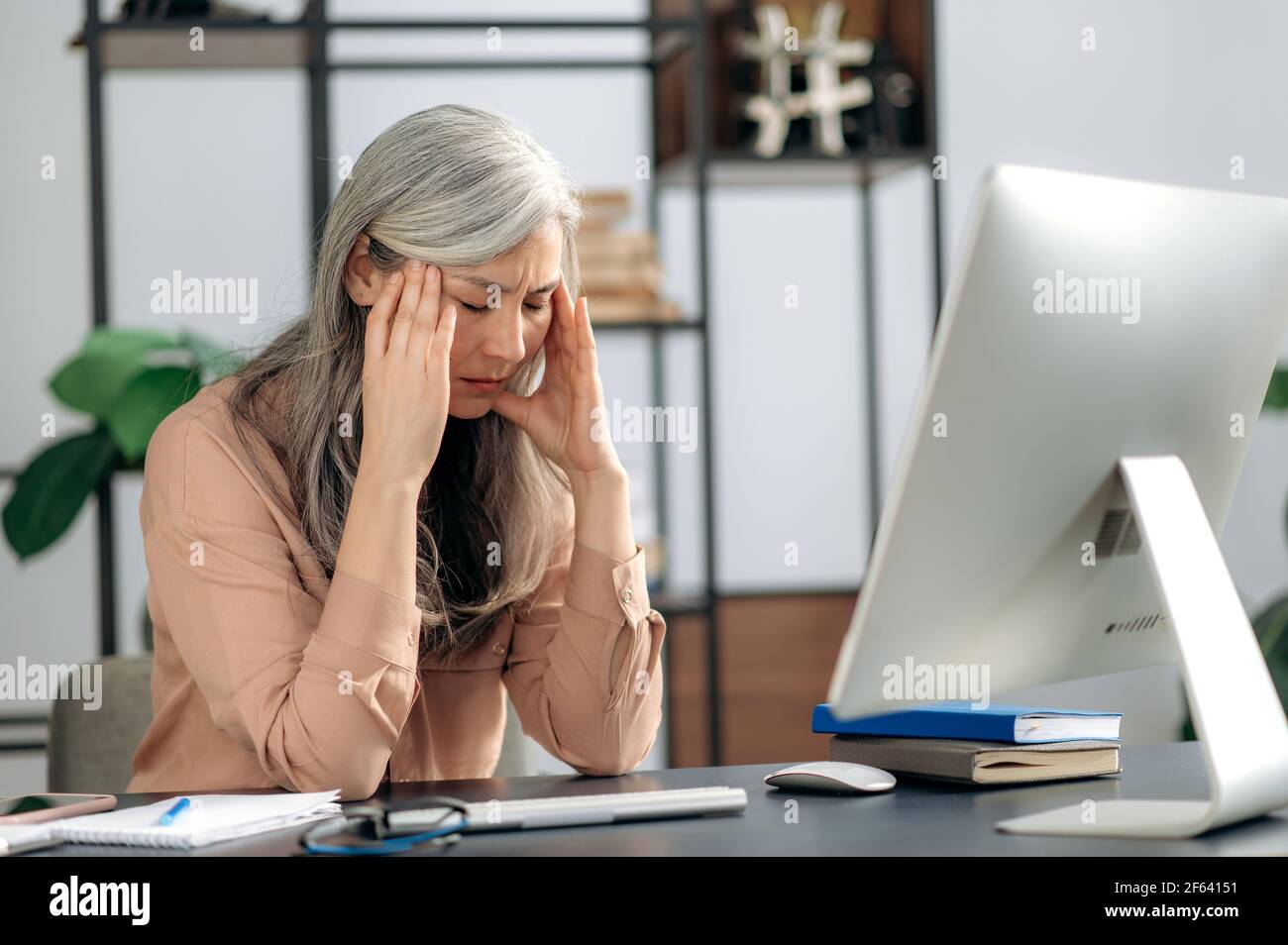 Concentrated tense senior gray-haired asian woman, manager, business lady, ceo, thinking about a project or deal, massaging her head with her hands, she has a headache, sitting at table, need rest Stock Photo