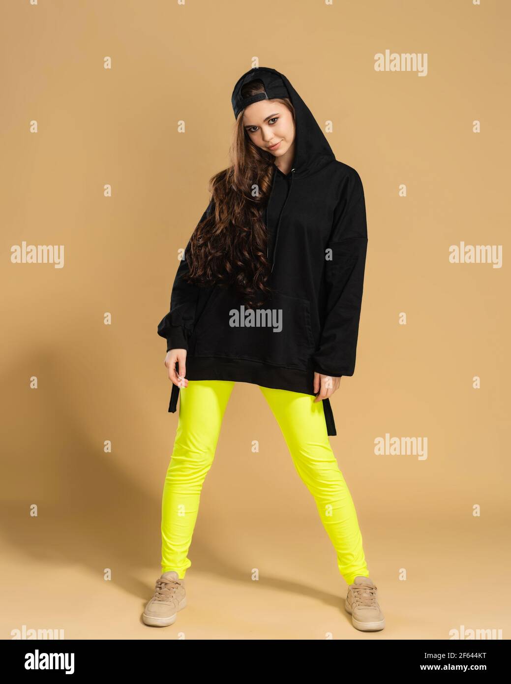 Young girl in yellow leggings, black hoodie and black baseball cap posing on a pastel orange studio background. Stock Photo