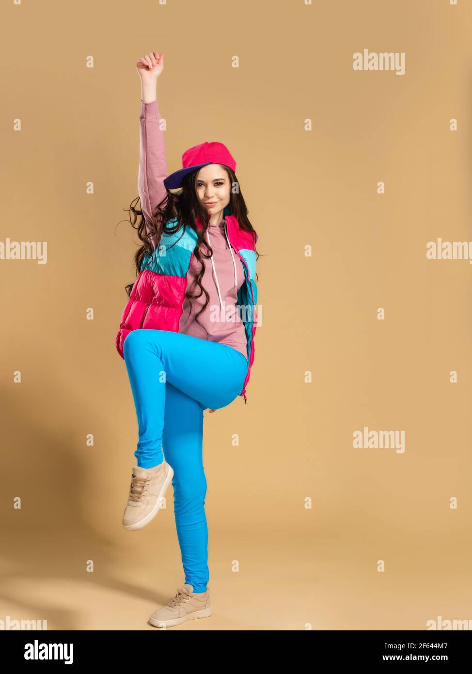 Young girl in a pink baseball cap with long hair in a bright multi-colored vest and blue leggings raises her hand up against an orange studio backgrou Stock Photo