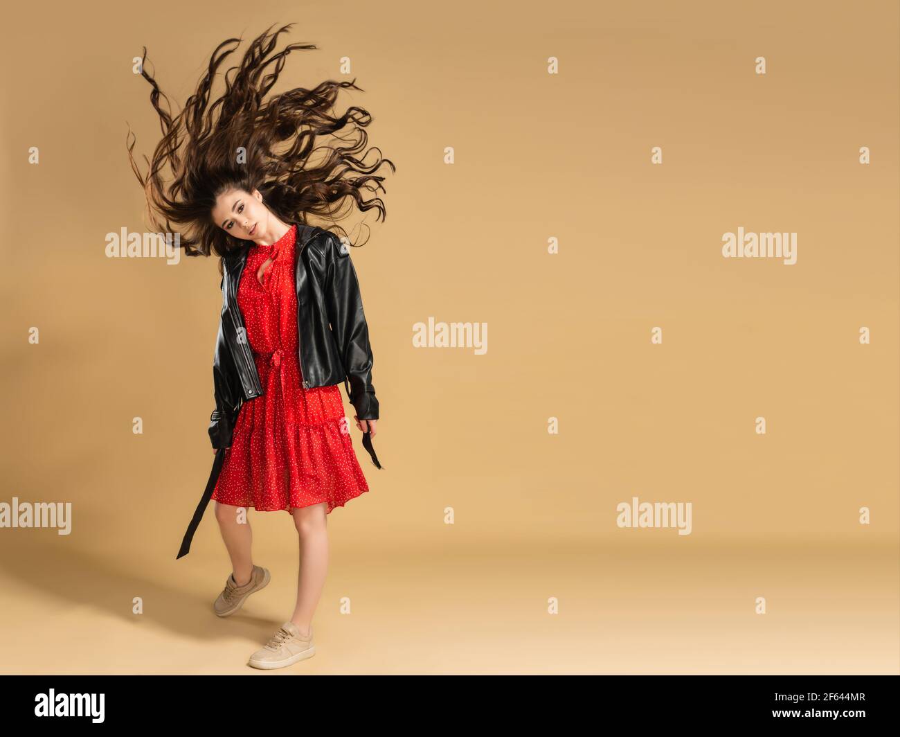 Young beautiful girl in a red dress with polka dots and a black leather jacket waving her long flowing hair on a pastel orange studio background. Stock Photo
