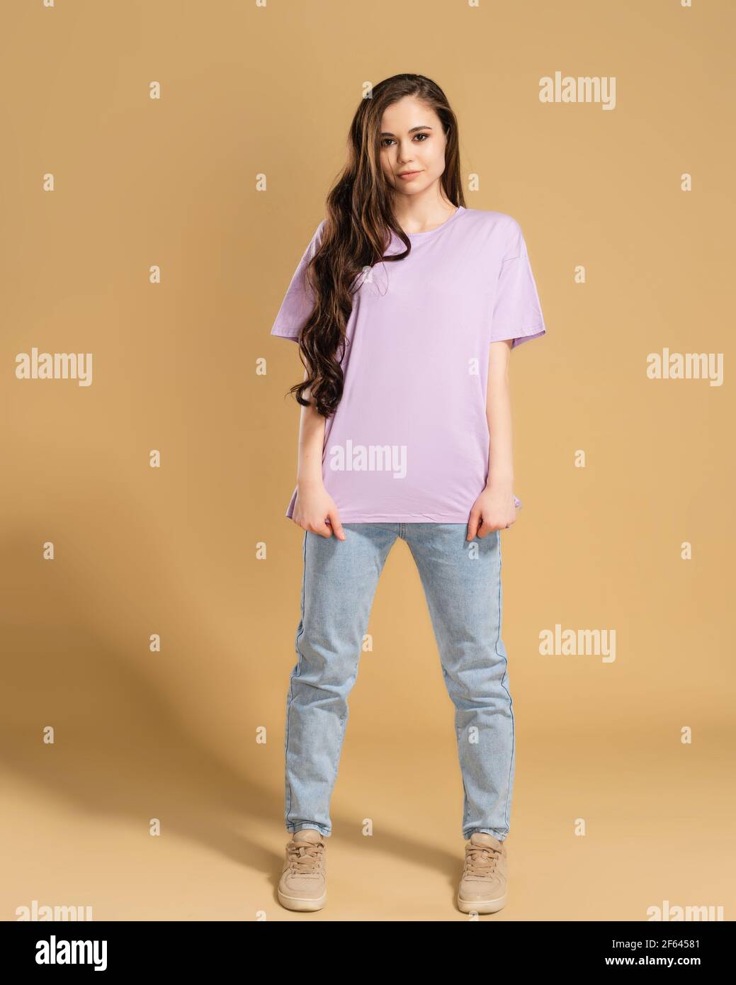 Young beautiful girl with long curly hair in a lilac T-shirt and blue jeans on a pastel orange studio background. T-shirt mockup. Stock Photo