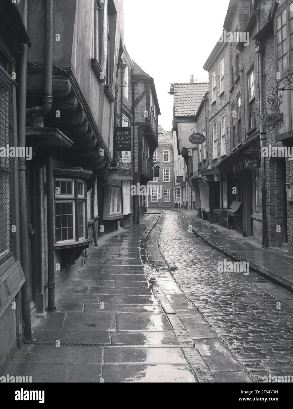 1956, historical, on a wet, damp British day, a view along an empty Shambles, a narrow, cobbled medieval street in York, England, UK, with overhanding timber-frame buildings, some dating as far back to the fourteenth century. Once known as 'The Great Flesh Shambles', from the meat that the butchers used to display in their shop windows, the ancient narrow street is perhaps Europe's best-preserved medieval street. The name 'Shambles' is now commonly used to collectively refer to the surounding maze of narrow twisting lanes and alleys as well. A sign for the York Weavers can be seen. Stock Photo