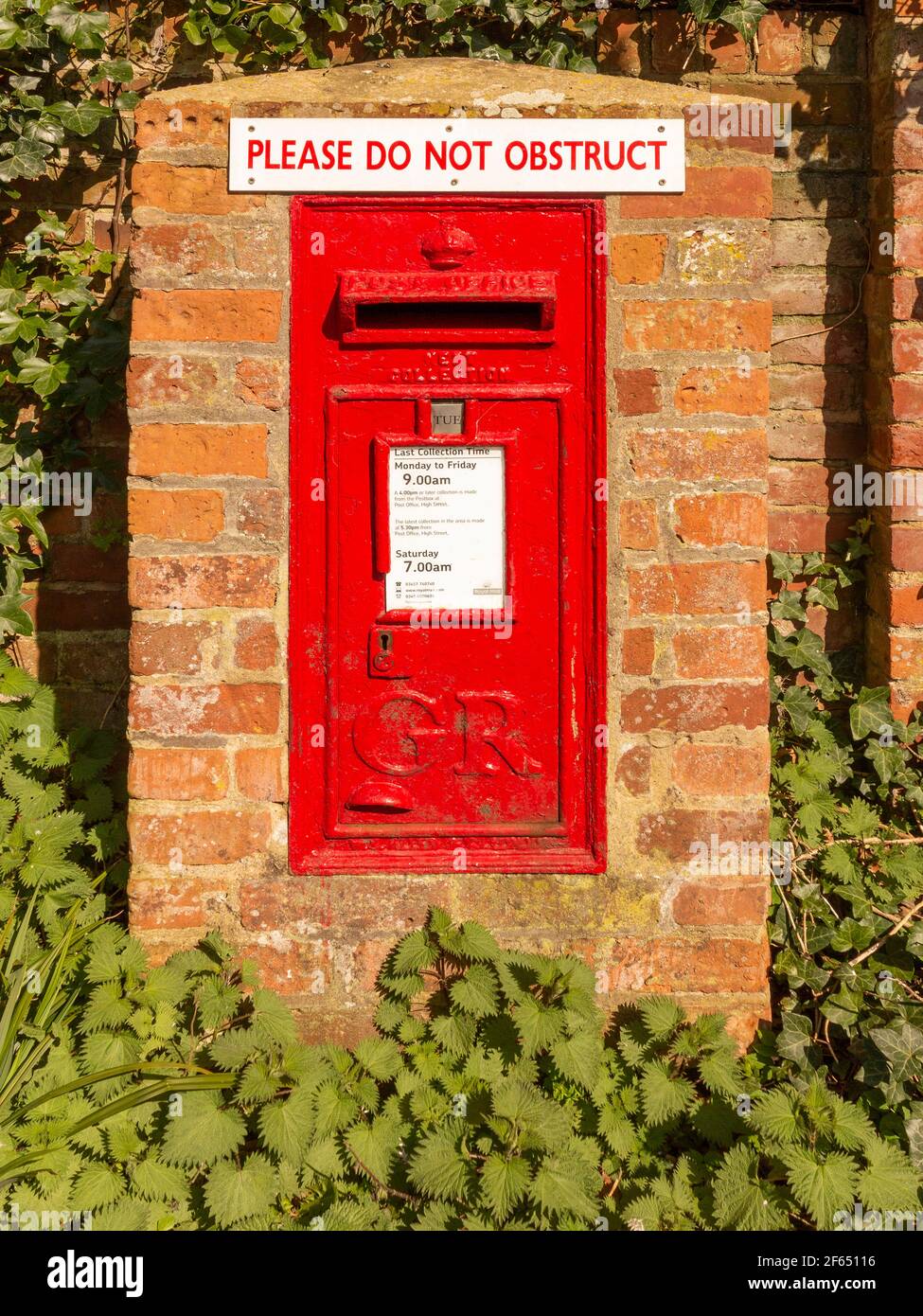 Wall mounted vintage post box from King George V era in its own brick structure surrounded by nettles and ivy with a 'Do not obstruct' sign attached. Stock Photo