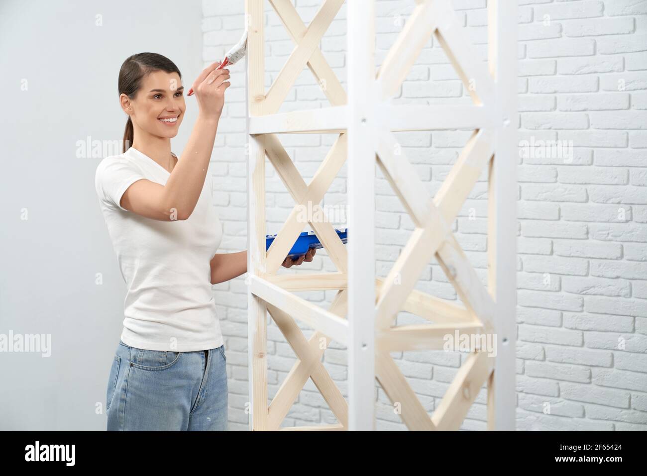 Smiling young brunette woman painting wooden rack at home. Concept of ...