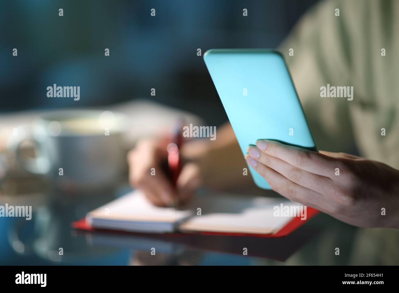 Close up of a woman hands checking smart phone calendar and taking notes in a paper agenda in the night at home Stock Photo
