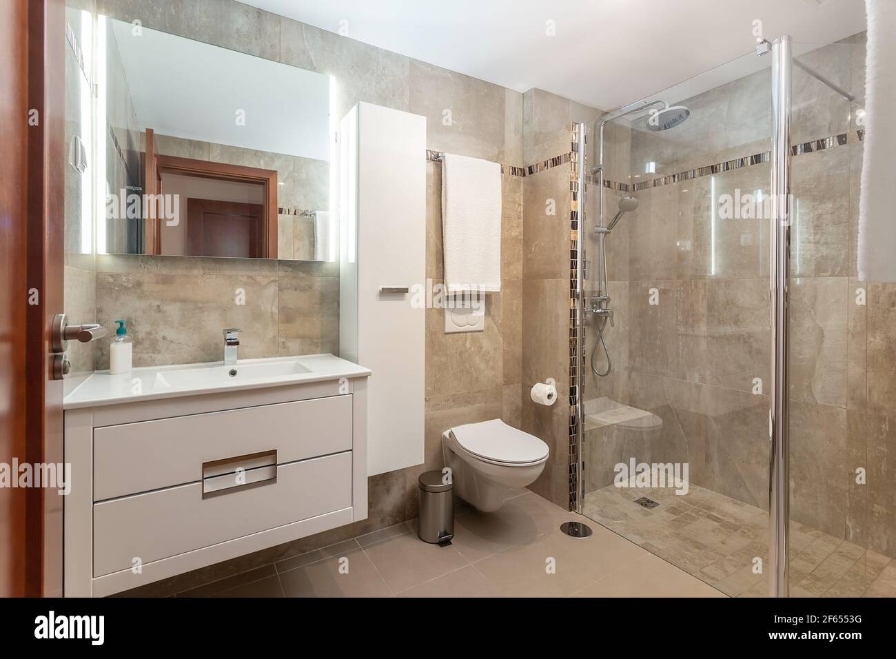 Beige colored bathroom interior, a shower with a glass door and a ceramic toilet, also a few towels on the walls. Stock Photo