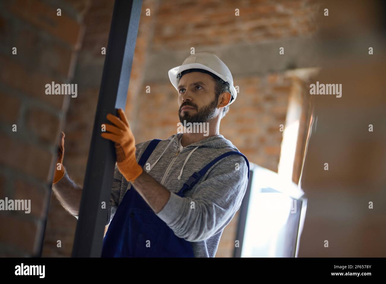 Focused young male builder wearing hard hat holding metal stud for drywall while working at construction site Stock Photo