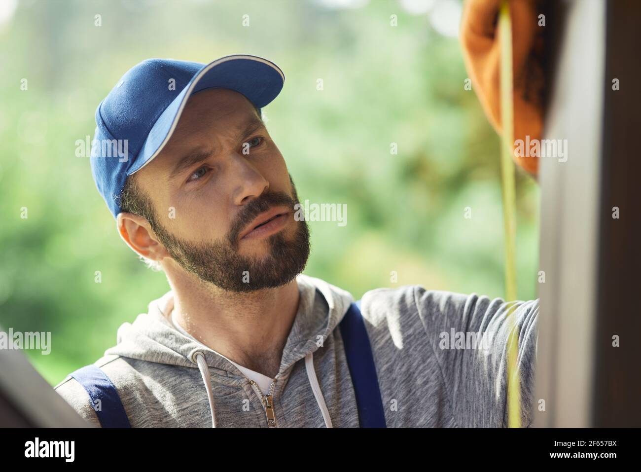 Close up portrait of focused young builder wearing blue cap and overalls using measuring tape while working on cottage construction on a sunny day Stock Photo