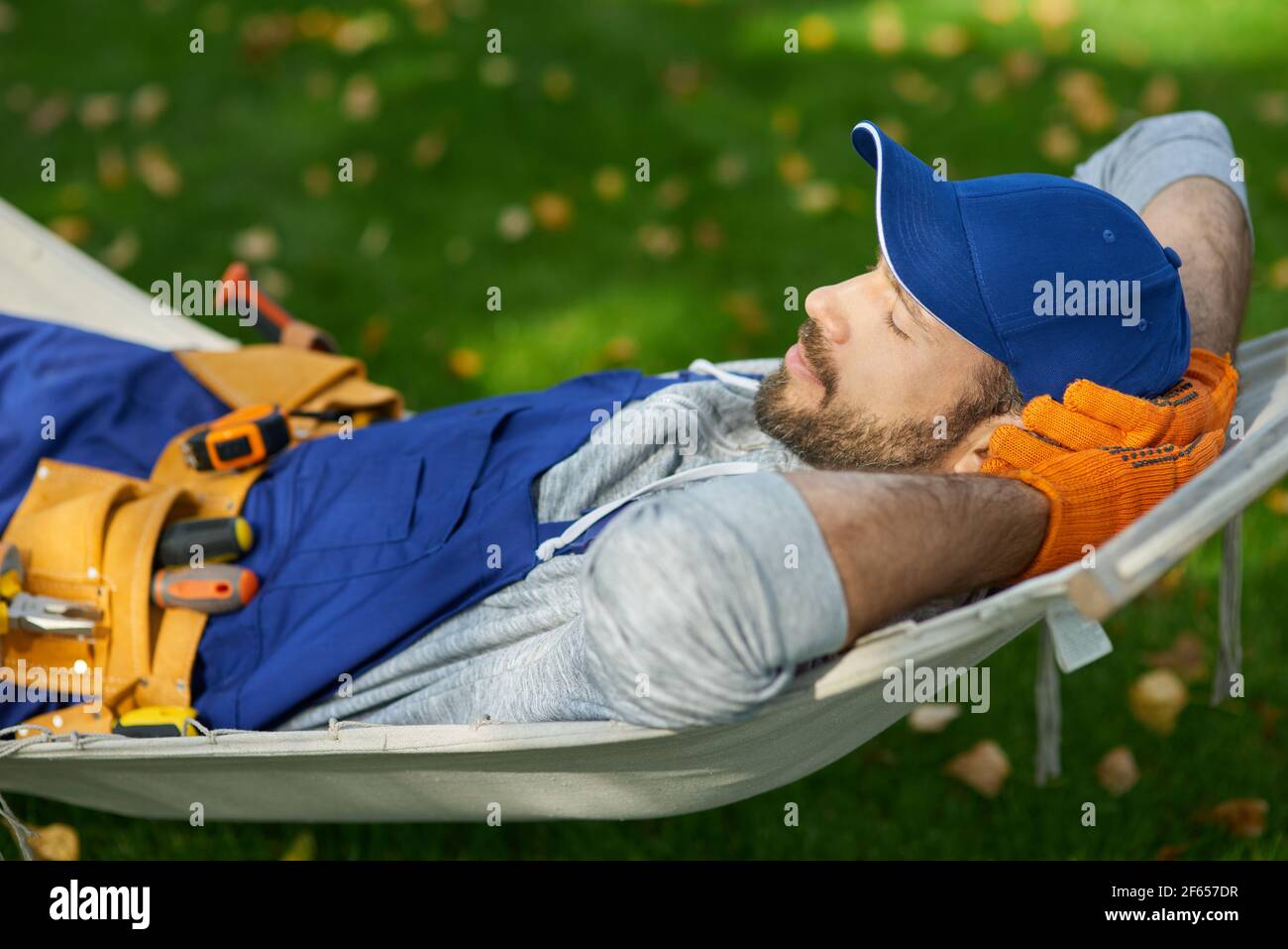 Peaceful young male builder wearing uniform taking a break, lying in a hammock outdoors with eyes closed on a sunny day Stock Photo