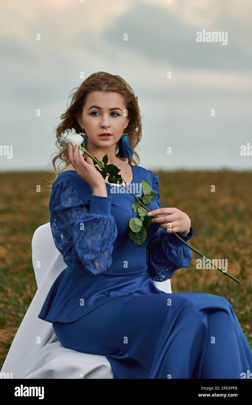 romantic woman portrait. female in blue dress holds white rose in her hands  while sitting on chair in the field Stock Photo - Alamy