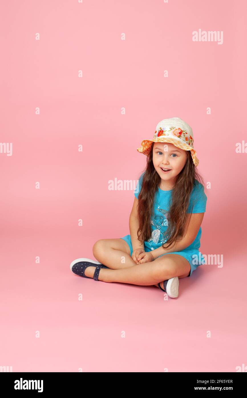 portrait of doubting, shocked girl with long wavy hair in a straw hat and blue dress sitting cross-legged on the floor, isolated on a pink background Stock Photo