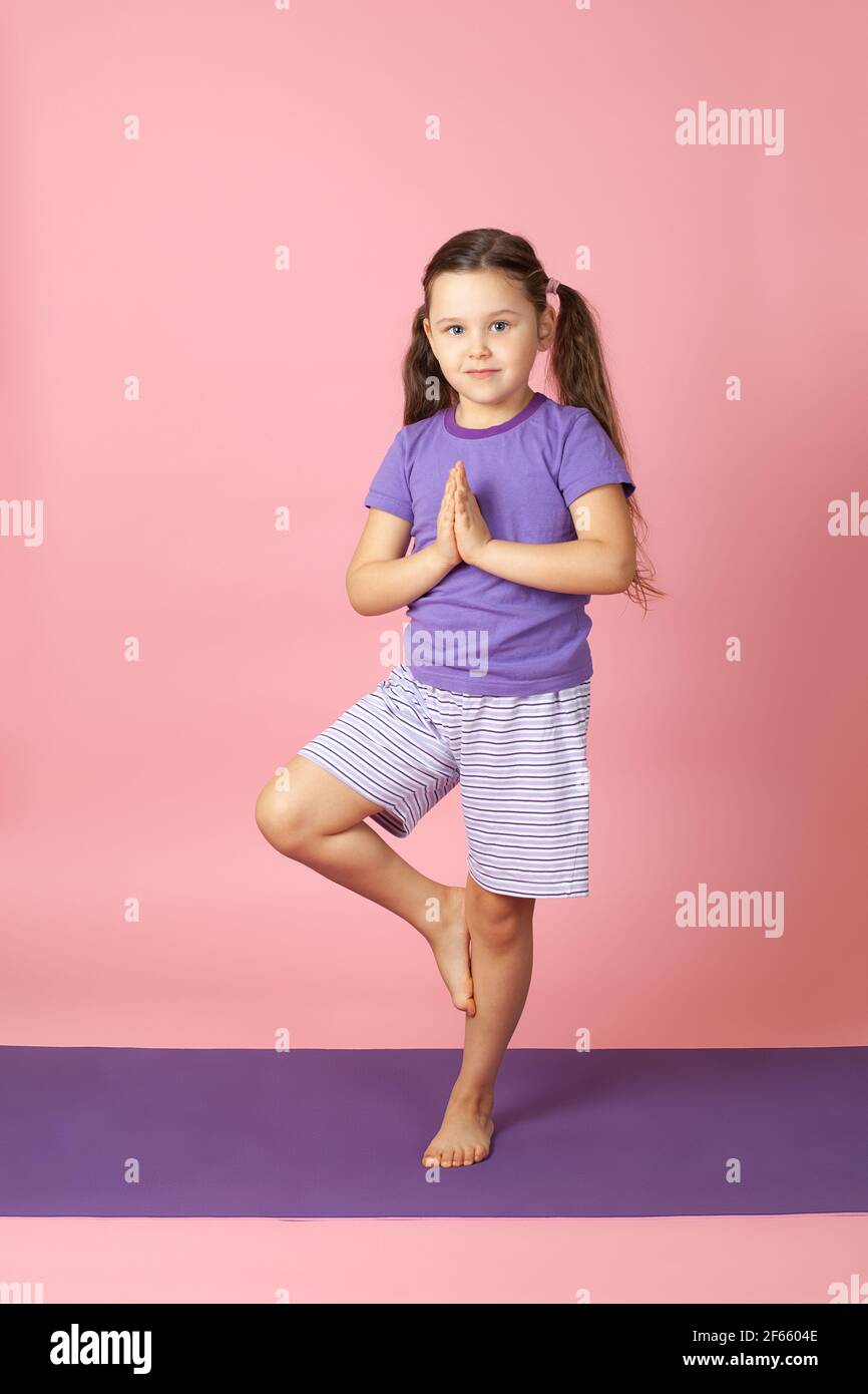 full-length portrait of a girl in shorts and a T-shirt on a purple mat in a tree pose in yoga, isolated on a pink background Stock Photo