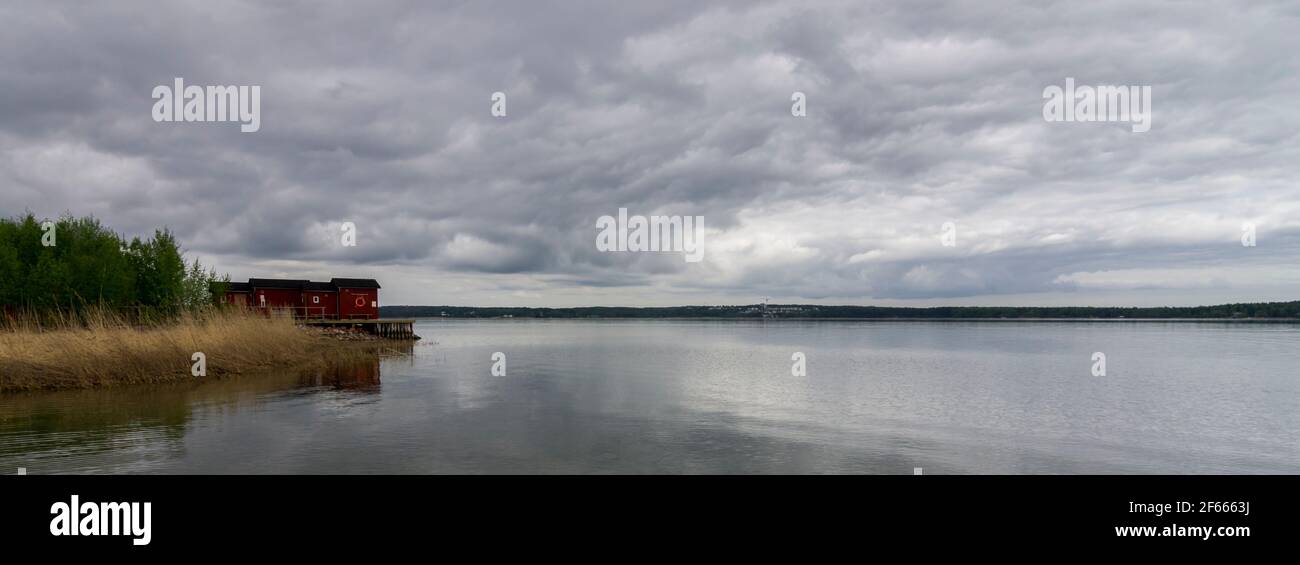 A red hut on the shore breaks up a view of grey, cloudy sky, and calm grey sea at an inlet near Mariehamn, Aland Islands, Finland. Stock Photo