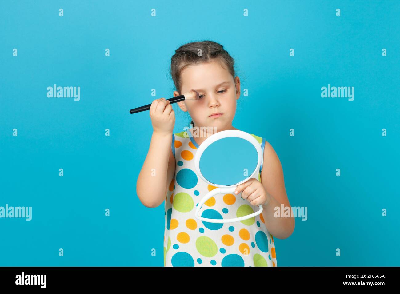 a pensive girl in a white dress powders her face, does makeup in front of a mirror in her hand, isolated on a blue background Stock Photo