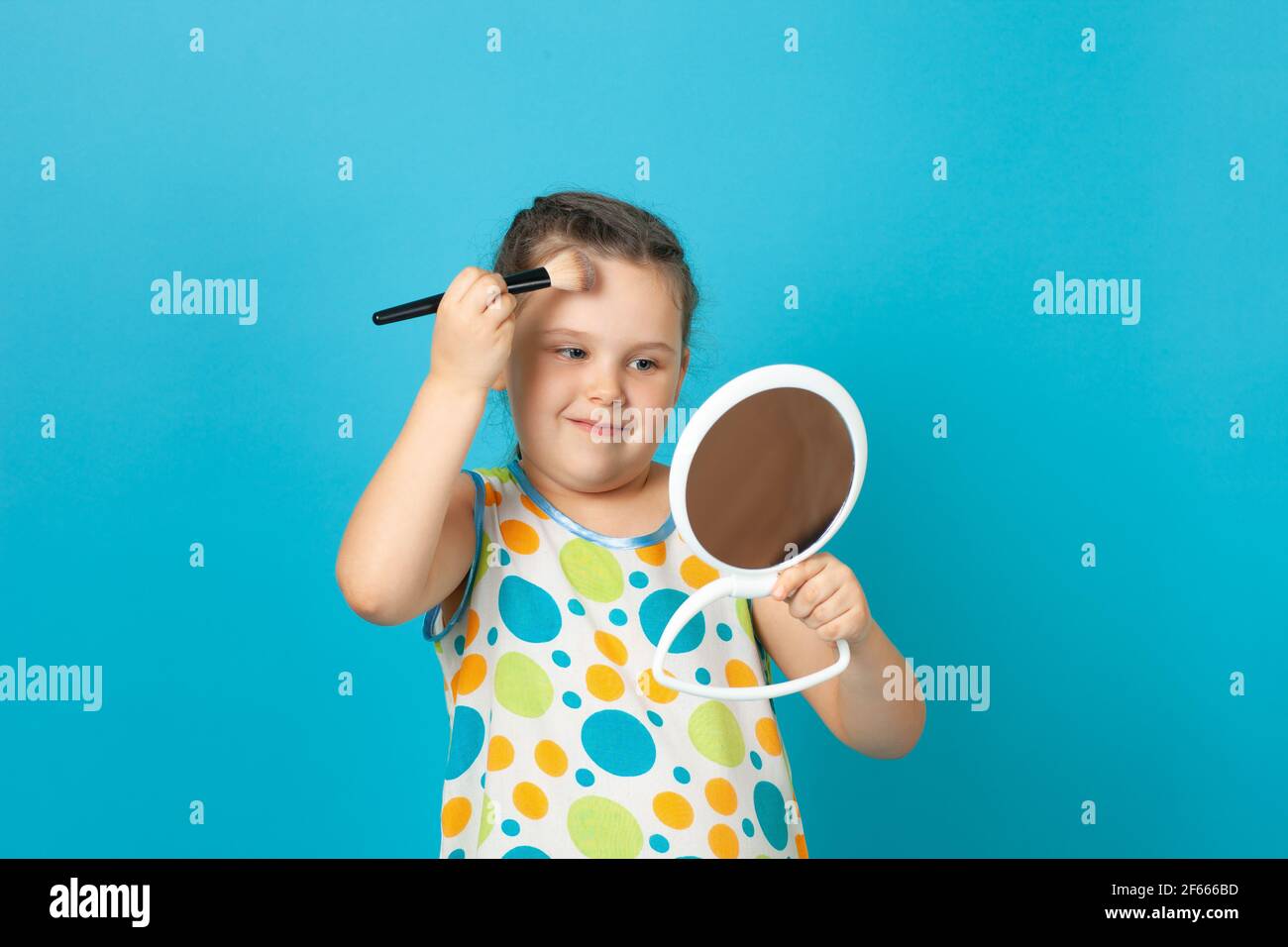 portrait of a girl in a white summer dress powdering her forehead with a large brush and doing blogger makeup, isolated on a blue background Stock Photo