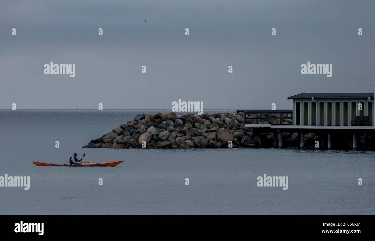 A kayaker floats past a stone pier / sea wall and a wooden hut on a gloomy evening in the Oresund Strait, Ribersborgsstranden, Malmo, Sweden. Stock Photo