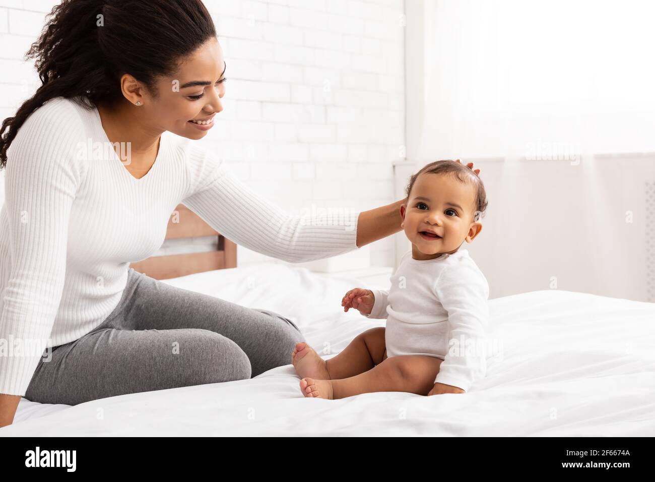 African American Mom Stroking Baby Toddler Sitting On Bed Indoor Stock Photo