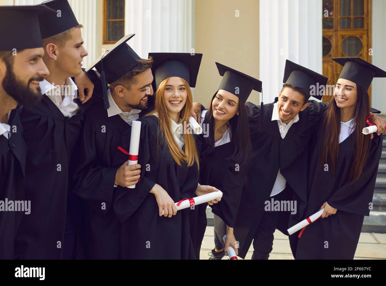 Smiling students in black gowns and square caps with diplomas having fun after graduation ceremony Stock Photo