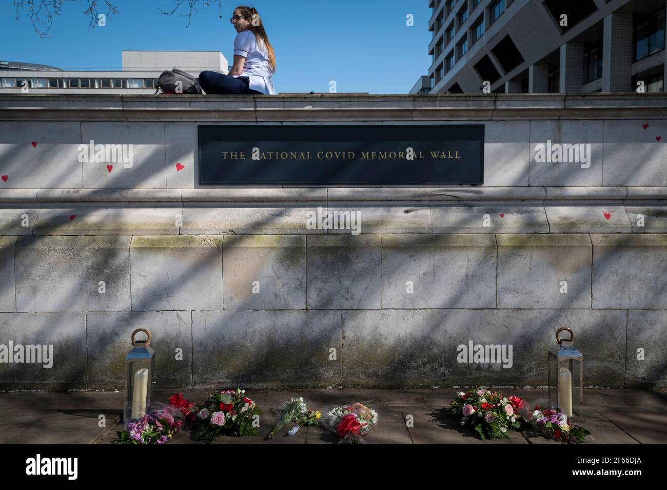 London, UK. 30th Mar, 2021. An NHS worker sits above the sign for hearts on a wall in Lambeth by the River Thames, with each heart representing someone who died during the UK's ongoing coronavirus pandemic. Called The National Covid Memorial Wall, it has been created by the Covid-19 Bereaved Families for Justice and will extend for half a mile by the time it is complete. Credit: Stephen Chung/Alamy Live News Stock Photo