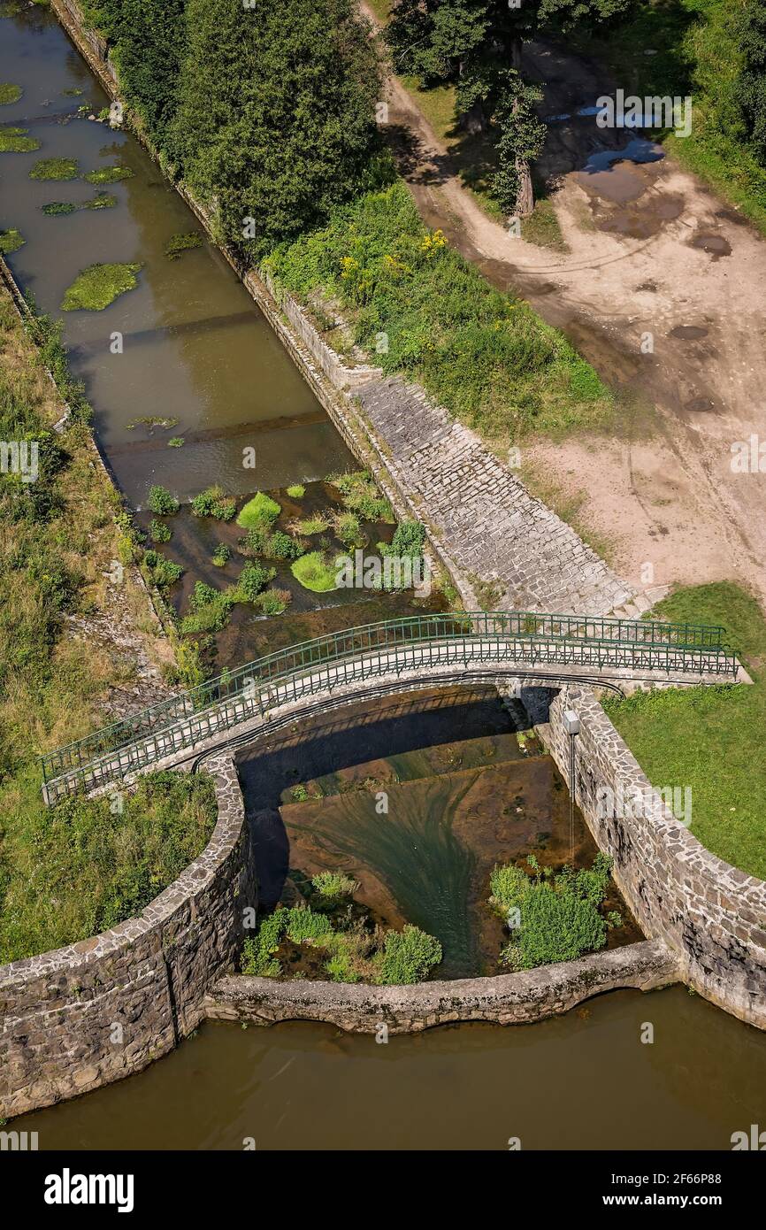 A vertical shot of a narrow river covered with green moss and plants and a tiny arched bridge Stock Photo