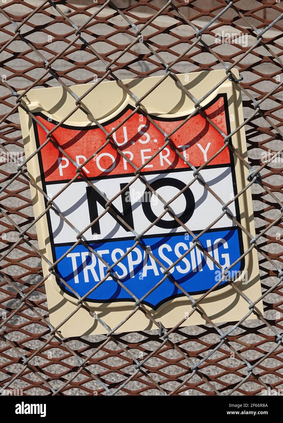 A sign reading U.S. PROPERTY NO TRESPASSING is shown between two metal security fences during the day in a vertical view. Stock Photo