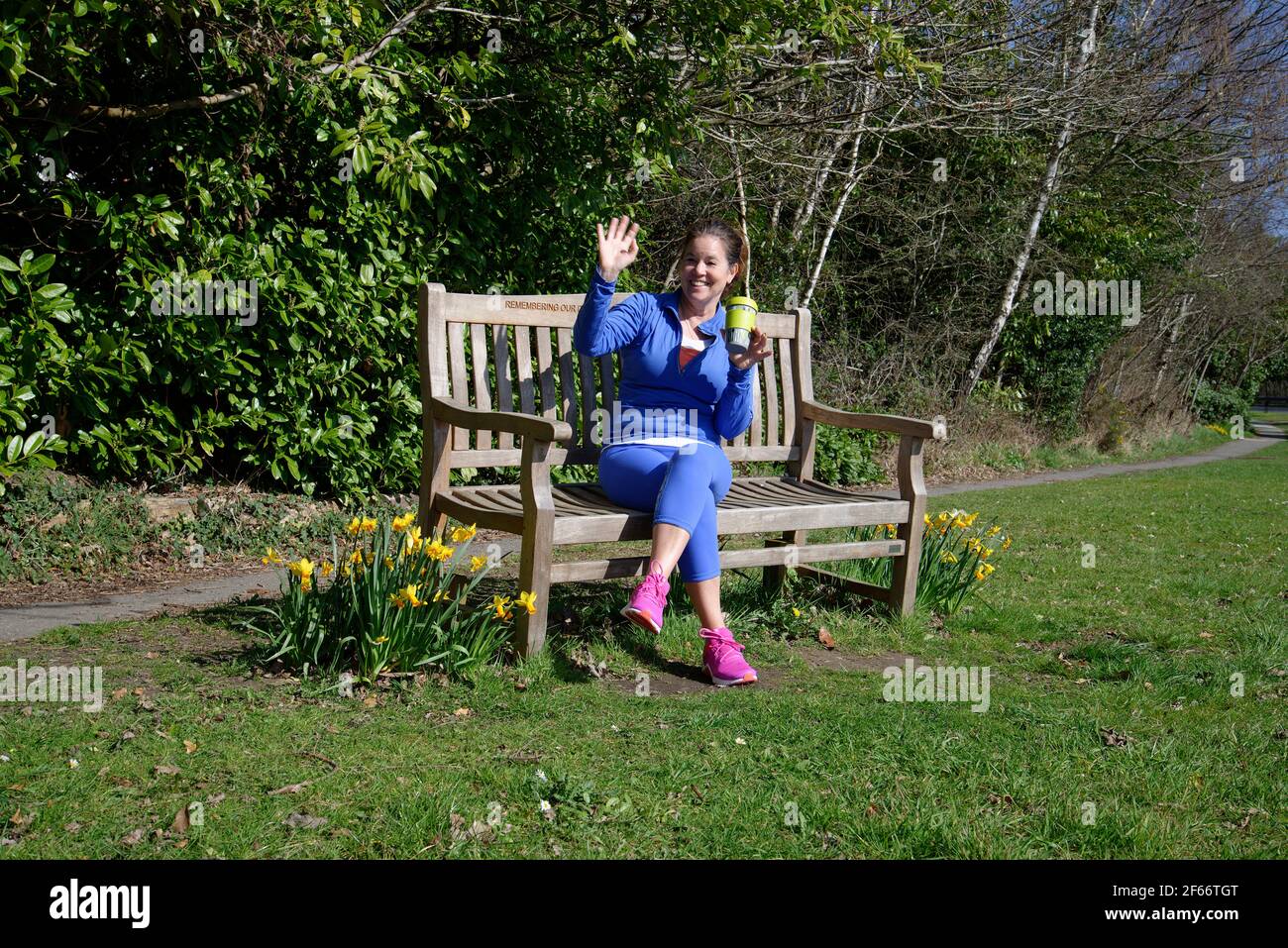 A middle aged woman sitting on a park bench after taking exercise. Cup of coffee in hand and waving to a friend. Stock Photo