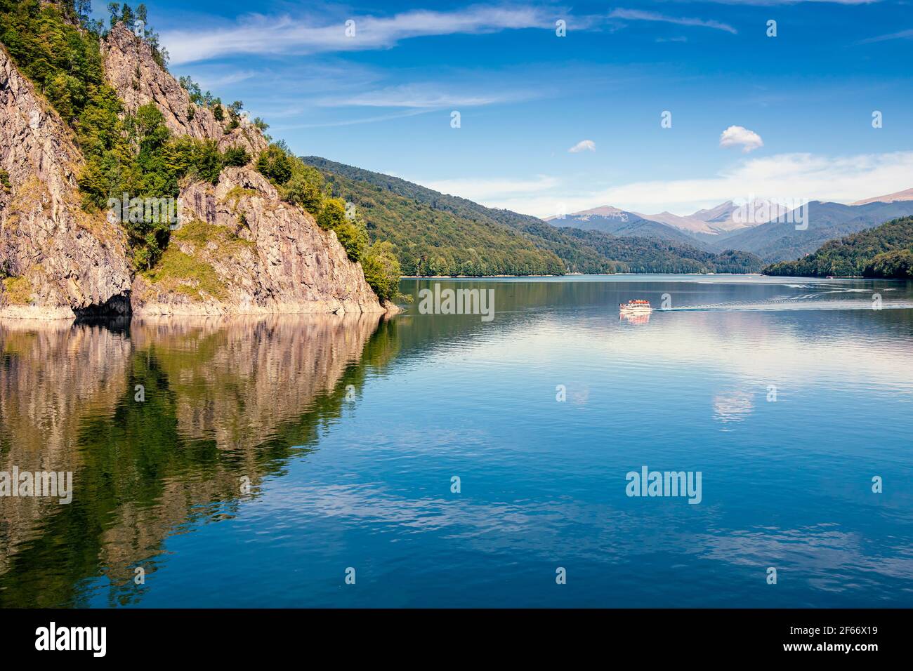 Daylight view to Vidraru lake in Carpathian Mountains. Bright blue sky and green trees. Boat cruising on water. Negative copy space, place for text. T Stock Photo