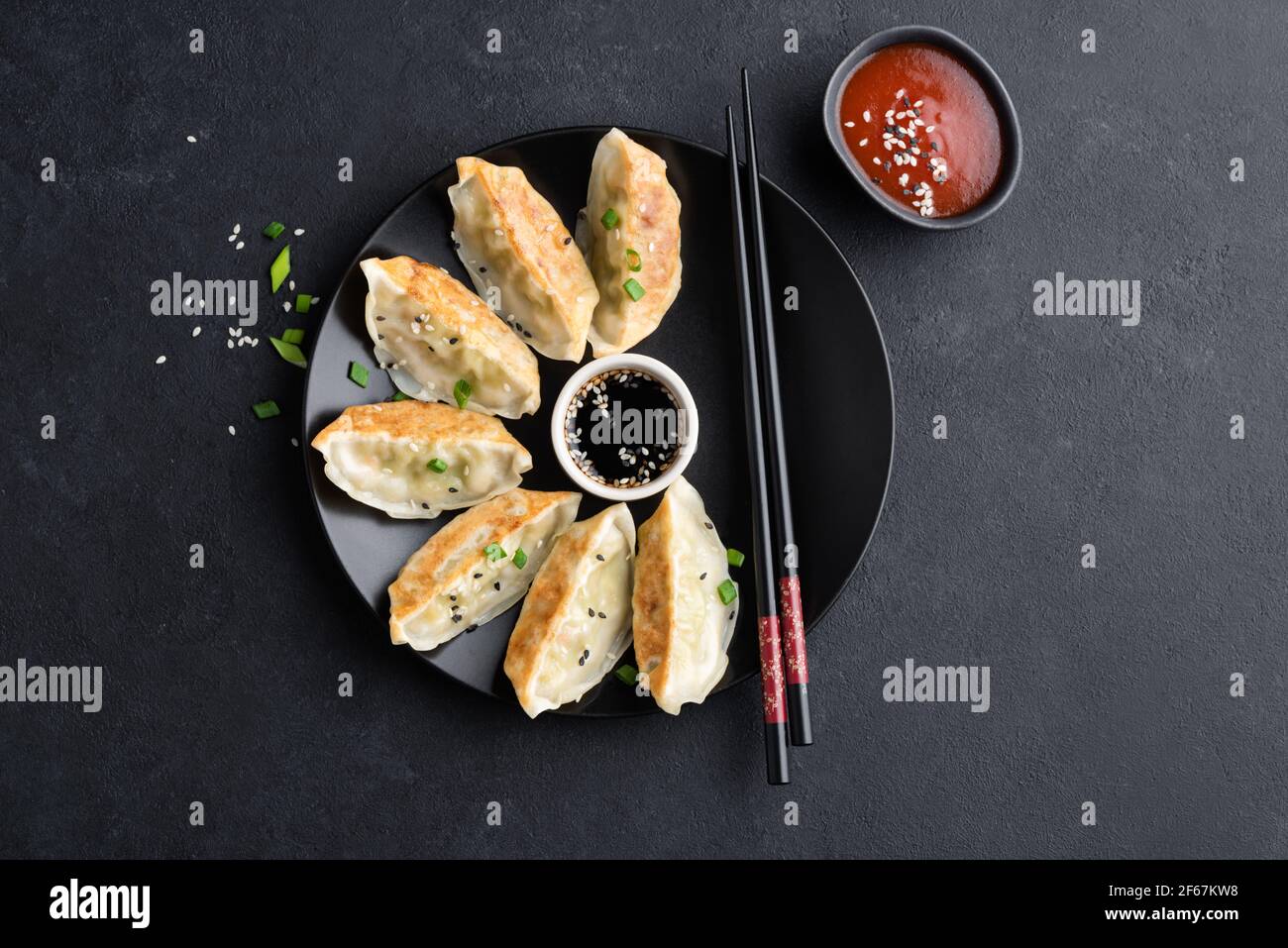 Asian food Gyoza or Jiaozi fried dumplings served with soy sauce, shriracha sauce and sesame seeds on black concrete background, top view Stock Photo
