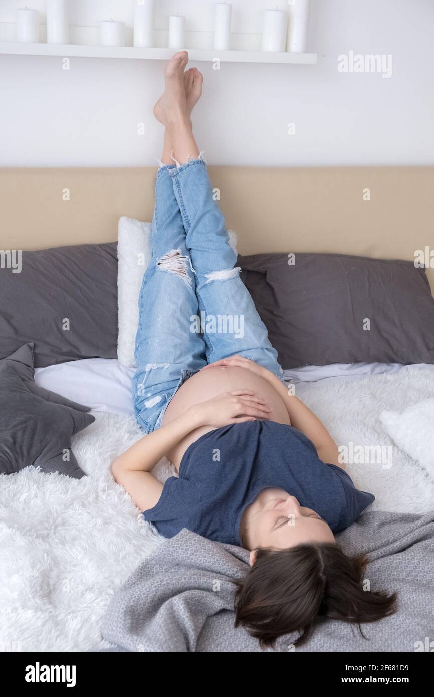 Pregnant young adult woman dressed jeans and t-shirt lying on bed relaxing with legs raised up Stock Photo