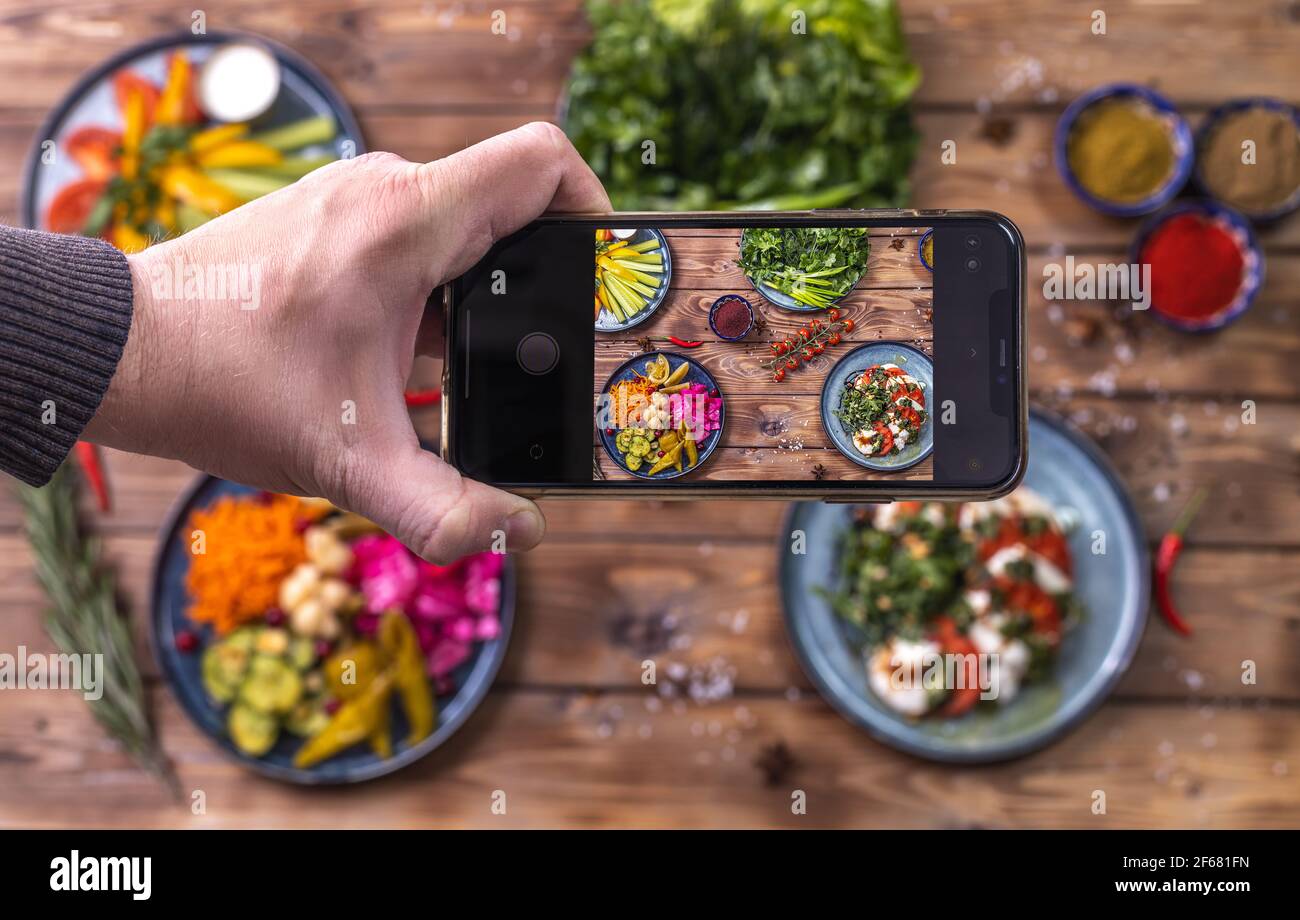 A person takes photos of pickled vegetables on his phone: peppers, carrots, garlic, cabbage on a wooden background decorated with chili peppers, cherr Stock Photo